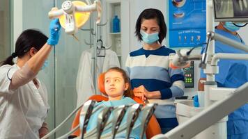 Orthodontist lighting the lamp until examining kid and patient opening mouth. Stomatologist speaking to mother of girl with toothache sitting on stomatological chair while nurse preparing tools. photo