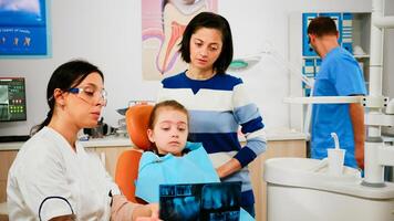 Pediatric dentist showing dental problems holding radiography pointing on affected teeth while man assistant preparing sterilized tools for surgery. Doctor and nurse working in stomatological unit photo