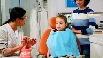 Pediatric stomatologist explaining to little patient procedure of extraction using model of dental teeth. Doctor holding sample of human jaw telling informations for keeping healthy teeth, digital x-ray in background photo