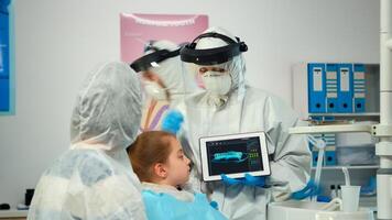Dentist with ppe suit pointing on digital screen explaining x-ray to mother of girl patient. Medical team and patients wearing face shield coverall, mask, gloves, showing radiography using notebook photo