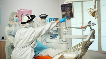 Dentist in ppe suit pointing on digital dental monitor explaining x-ray to mother and kid wearing coverall during global pandemic. New normal dentist visit in coronavirus outbreak with face shield photo