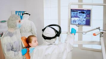 Stomatologist in coverall explaining dental problem using digital monitor during global pandemic. Assistant and doctor with coverall face shield mask gloves examining woman from stomatological chair photo