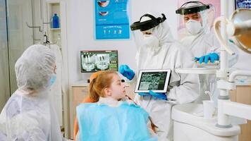Stomatologist in protective equipment showing on tablet dental x-ray reviewing it with mother of patient. Medical team wearing face shield mask, gloves, explaining radiography using notebook display photo