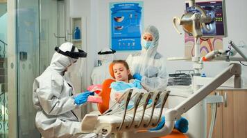 Stomatologist in ppe suit holding plaster model of the mandible speaking with girl patient. Medical team wearing face shield coverall, mask, gloves, showing correct dental hygiene using teeth skeleton photo