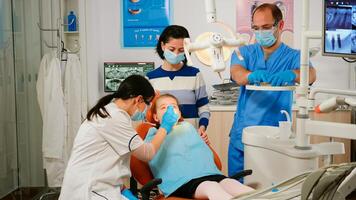 Woman dentist technician lighting the lamp for examining little patient. Orthodontist speaking to girl sitting on stomatological chair while nurse preparing tools for examination in modern clinic. photo