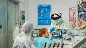 Dentist in protective equipment showing on tablet dental x-ray reviewing it with mother of kid patient. Medical team wearing face shield coverall, mask, gloves, explaining radiography using notebook photo