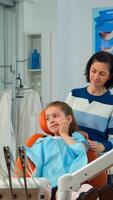 Pediatric woman dentist talking with mother and girl about eating habit and oral hygiene of the child before dental examination. Sad kid patient is showing sick tooth while nurse preparing dental tools photo