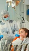 Dentist doctor in ppe suit interrogating kid patient and taking notes on clipboard while girl indicating affected mass. Stomatologist and assistent working in new normal dental office wearing coverall photo