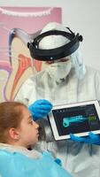 Dentist with ppe suit pointing on digital screen explaining x-ray to mother of girl patient. Medical team and patients wearing face shield coverall, mask, gloves, showing radiography using notebook photo