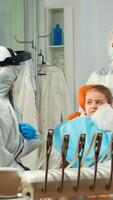 Child in protection suit showing dental problem to orthodontist wearing coverall during global pandemic. Concept of new normal dentist visit in coronavirus outbreak wearing face shield photo