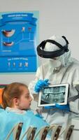 Dentist in protective equipment showing on tablet dental x-ray reviewing it with mother of kid patient. Medical team wearing face shield coverall, mask, gloves, explaining radiography using notebook photo