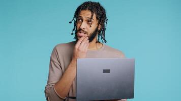 Middle Eastern man inputting data on laptop while standing in blue studio background. BIPOC employee typing on notebook keyboard, focused on solving project tasks, camera B photo