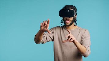 Tech enthusiast wearing virtual reality goggles, doing swiping gestures. BIPOC man using high tech futuristic modern VR glasses, making tactile touching gesturing, blue studio background photo