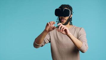Middle Eastern man wearing virtual reality headset, doing swiping gestures. BIPOC person using high tech futuristic modern VR gadget, isolated over blue studio background, camera A photo