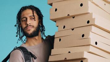 Portrait of smiling pizza delivery man holding takeaway orders for customer. Upbeat BIPOC courier with lunch boxes in arms and thermal food backpack, isolated over blue studio background, camera B photo