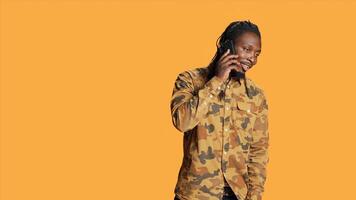 Young adult chatting on phone call in studio, talking to people on remote telephone line over orange background. African american person having fun with telecommunication conversation. photo
