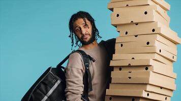 Portrait of smiling pizza delivery man holding takeaway orders for customer. Upbeat BIPOC courier with lunch boxes in arms and thermal food backpack, isolated over blue studio background, camera A photo