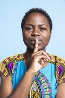 African american girl doing silence privacy symbol in studio, showing mute gesture to keep secret and be private. Young silent person doing hush sign over blue background, confidential information. photo