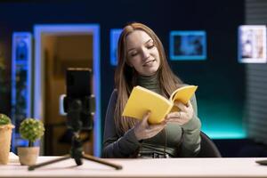 Influencer in studio using camera to capture live broadcast, reviewing new book from publisher. Online show host filming literature critique, presenting new novel from esteemed author to viewers photo