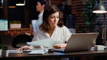 Worker looking over paperwork containing financial graphs, researching key data for company project. Businesswoman looking over accounting figures and charts on papers late at night at desk photo