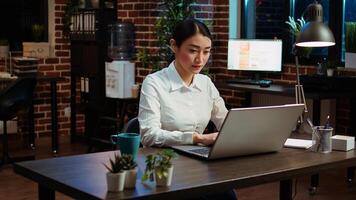 Smiling woman sitting down at office desk, typing on laptop keyboard, starting work with mug full of coffee, solving tasks late at night. Cheerful employee imputing data on computer photo