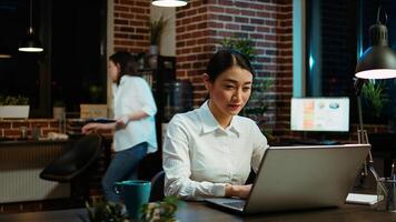 Portrait of smiling asian businesswoman doing computer tasks for team project in office. Cheerful employee working late at night, typing on laptop keyboard next to coworker, camera A photo