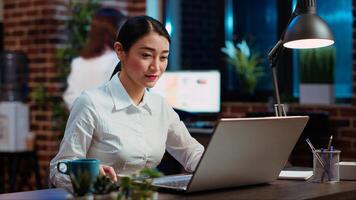 Businesswoman drinking cup of coffee while doing tasks in office overnight for company project. Employee enjoying hot beverage while looking over figures on laptop late at night, typing on keyboard photo