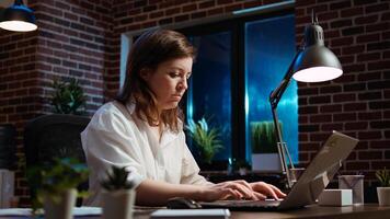 Businesswoman researching key data for company project, doing tasks in office overnight. Employee looking over accounting figures on laptop screen late at night, typing on keyboard, camera A photo