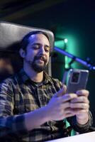 Cheerful male student is utilizing his mobile device to browse social media while seated at his desk. Bearded man with a smile answers texts while using his smartphone for surfing the internet. photo