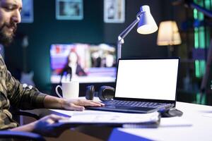 Male freelancer working from home utilizing portable computer with a blank chromakey mockup template. Bearded man looking at his clipboard while his digital laptop displays empty white screen photo