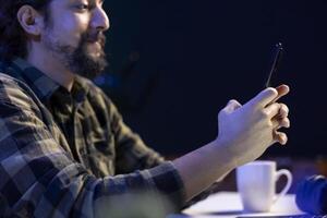 Cheerful male student is utilizing his mobile device to check social media while seated at his desk. Bearded man with a smile answers texts while using his smartphone to browse the internet. photo