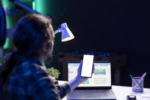 Bearded man vertically holding mobile device displaying isolated white screen. Male freelancer working on his laptop while grasping smartphone with blank copyspace mockup template. photo