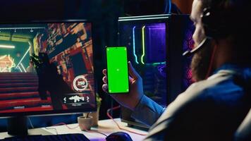 Man watching tutorial on green screen phone while playing first person shooter videogame with gun shooting laser bullets. Gamer learning to play game by looking at online guide on smartphone photo
