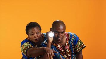 Confident man using flashlight to see what caused noise, trying to calm his wife who is hiding behind him. Terrified woman being anxious about weird sound, husband using lamp light and searching. photo