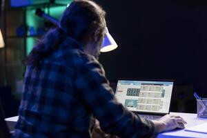 Serious man sits at his desk, closely inspecting stock information on laptop. Male freelancer focused on device screen, which displays a forex trading chart and analyzing the value of economic trends. photo