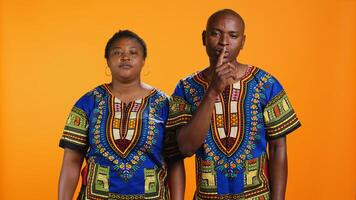 Ethnic man and woman showing hush privacy sign on camera, putting finger over lips to keep secret over orange background. Young couple presenting mute gesture, asking for secrecy. photo