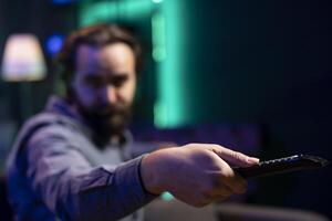 Focus on remote control used by man in blurry background switching channels on television screen. Close up of device used by person to scroll through streaming content, picking movie photo