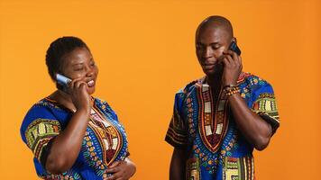 Cheerful couple talking to family on phone call, having remote conversation with smartphone over orange background. Young people in traditional clothes chatting on telephone line. photo