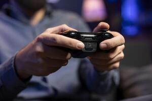 Focus shot on controller held by gamer in blurry background playing videogames. Close up shot of gamepad used by man enjoying gaming session in living room while sitting down in front of TV photo