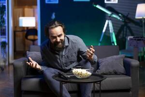 Happy man laughing and enjoying bowl of popcorn, watching comedy cable programs on TV during relaxation time at home. Cheerful spectator on couch amused by funny television series photo