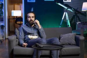 Man watching shows on TV and eating popcorn, engrossed by jaw dropping action, paying close attention to screen. Man sitting on couch in apartment eating popcorn, shocked by wild film plot photo