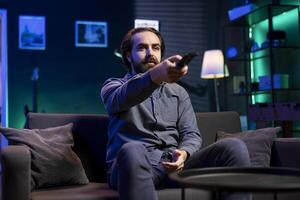 Man watching shows on TV, using remote control, paying close attention to screen. Sedentary man sitting in pajamas all day consuming mindless entertainment, picking what movie to watch next photo