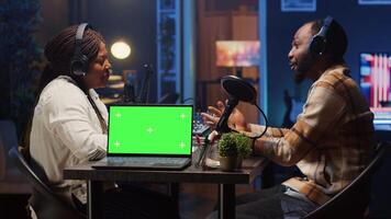 Isolated screen laptop next to vlogger show presenter and guest talking in neon lights ornate studio. Chroma key notebook next to man and woman broadcasting discussion on online podcast photo