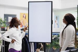 Shopping mall worker explaining shoes brand features to client using blank smart display. African american store assistant showcasing new collection to buyer on digital empty screen in boutique photo