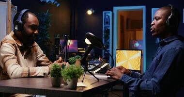Vlogger show host interviewing guest in neon lights ornate personal studio, discussion about his life. BIPOC men filming online podcast episode, talking with each other, jib down shot photo