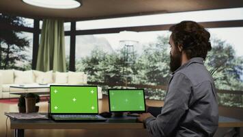 Businessperson using greenscreen displays on devices, sitting at his desk in luxurious villa in a forest. Rich entrepreneur looking at screens showing isolated copyspace, works on laptop and tablet. photo
