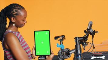 Woman holds green screen tablet, using it to advertise bike repair shop, empty placeholder for company logo, studio background. Mechanic promoting bicycle fixing service with mockup device, camera A photo