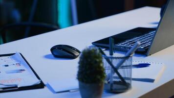Office desk filled with productivity items in empty home interior illuminated with neon lights at night. Laptop on table and other work related objects in apartment room, camera A close up shot photo