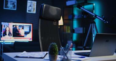 Apartment illuminated by RGB lights with office desk in front of smart TV displaying news program broadcasting. Laptop, mini house plants and headphones on table in empty room, camera B close up shot photo