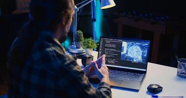 Computer scientist updating artificial intelligence machine learning algorithm, writing binary code scripts. Teleworking IT specialist in apartment office uses programming to upgrade AI, camera B photo